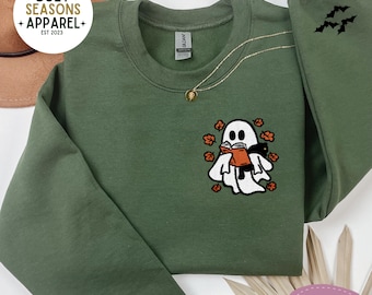 May include: Green sweatshirt with an embroidered ghost reading a book, surrounded by fall leaves. The sleeve features a bat design. The text "Cozy Seasons Apparel" is in the upper left corner.