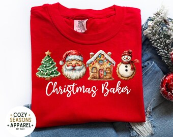 May include: A red t-shirt with a Christmas-themed design. It features a Christmas tree, Santa Claus, a gingerbread house, and a snowman. The words "Christmas Baker" are written in white script. The shirt has a crew neck and is made of a soft material.