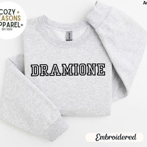 May include: A grey sweatshirt with the text "DRAMIONE" embroidered in black on the front. The sweatshirt is folded in half and the text "Embroidered" is visible below the sweatshirt.