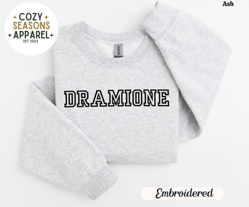 May include: A grey sweatshirt with the text "DRAMIONE" embroidered in black on the front. The sweatshirt is folded in half and the text "Embroidered" is visible below the sweatshirt.