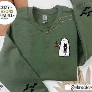 May include: A green sweatshirt with embroidered bats on the sleeves and a ghost holding a cat and a cup of coffee on the front. The text "Cozy Seasons Apparel Est 2023" is on the top left corner of the sweatshirt. The sweatshirt is labeled "Military Green" and "Embroidered".