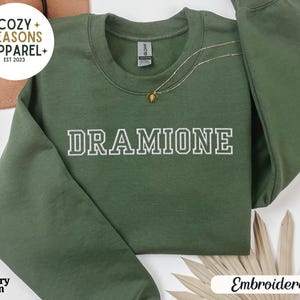 May include: A green sweatshirt with the text "Cozy Seasons Apparel Est 2023" in white on the top left corner. The text "DRAMIONE" is embroidered in white on the front of the sweatshirt.