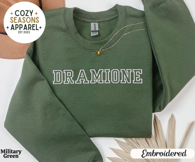 May include: A green sweatshirt with the text "Cozy Seasons Apparel Est 2023" in white on the top left corner. The text "DRAMIONE" is embroidered in white on the front of the sweatshirt.