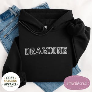 May include: Black hooded sweatshirt with the text "DRAMIONE" embroidered in white on the front. The sweatshirt is on a blue denim background.