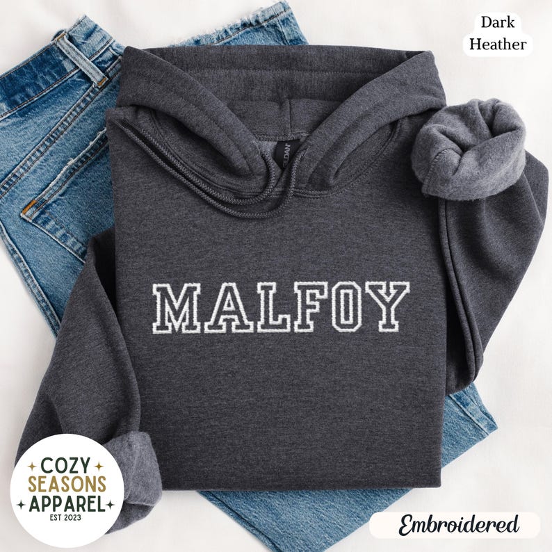 Peut inclure: Un sweat &agrave; capuche gris fonc&eacute; avec le mot "MALFOY" brod&eacute; en blanc. Le sweat &agrave; capuche a une capuche &agrave; cordon et des poignets c&ocirc;tel&eacute;s. L'image comprend &eacute;galement le texte "Cozy Seasons Apparel" et "Embroidered".