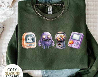 May include: Dark green sweatshirt featuring a row of whimsical watercolor illustrations: a retro TV with a ghost, a purple monster, a gumball machine, and a purple handheld game console with a pumpkin. The text "COZY SEASONS + APPAREL + EST 2023" is at the bottom.