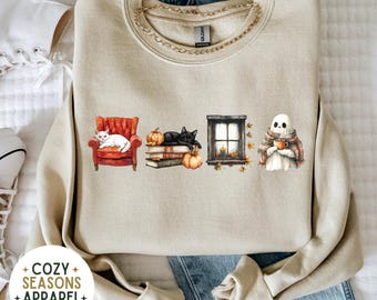 May include: Beige sweatshirt with a gold chain and a graphic of a white cat in a red chair, a black cat on books with pumpkins, a window, and a ghost with a cup. The text "COZY SEASONS APPAREL" is also visible.
