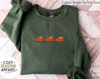 Può includere: Una felpa girocollo verde militare con un ricamo raffigurante tre fette di torta di zucca arancioni con ciuffi di panna montata bianca. Il capo presenta maniche lunghe con polsini a coste e un colletto a coste dal design semplice e pulito.