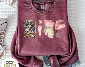 May include: A maroon sweatshirt with a graphic design of baking supplies, including a muffin tin, piping bag, oven mitt, and a stand mixer. The text "Bake" is spelled out using the baking supplies.