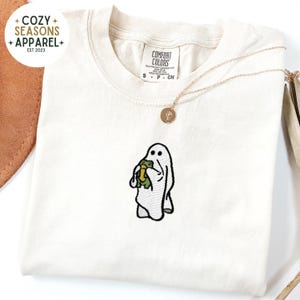 May include: A white t-shirt with an embroidered ghost holding a frog. The shirt has the text "COMFORT COLORS" on the tag.
