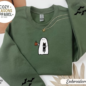 May include: A green sweatshirt with an embroidered design of a ghost holding a cat. The ghost is holding a cup of coffee. The sweatshirt has the text "Cozy Seasons Apparel" and "Est 2023" on the top left corner. The sleeves have embroidered bats.