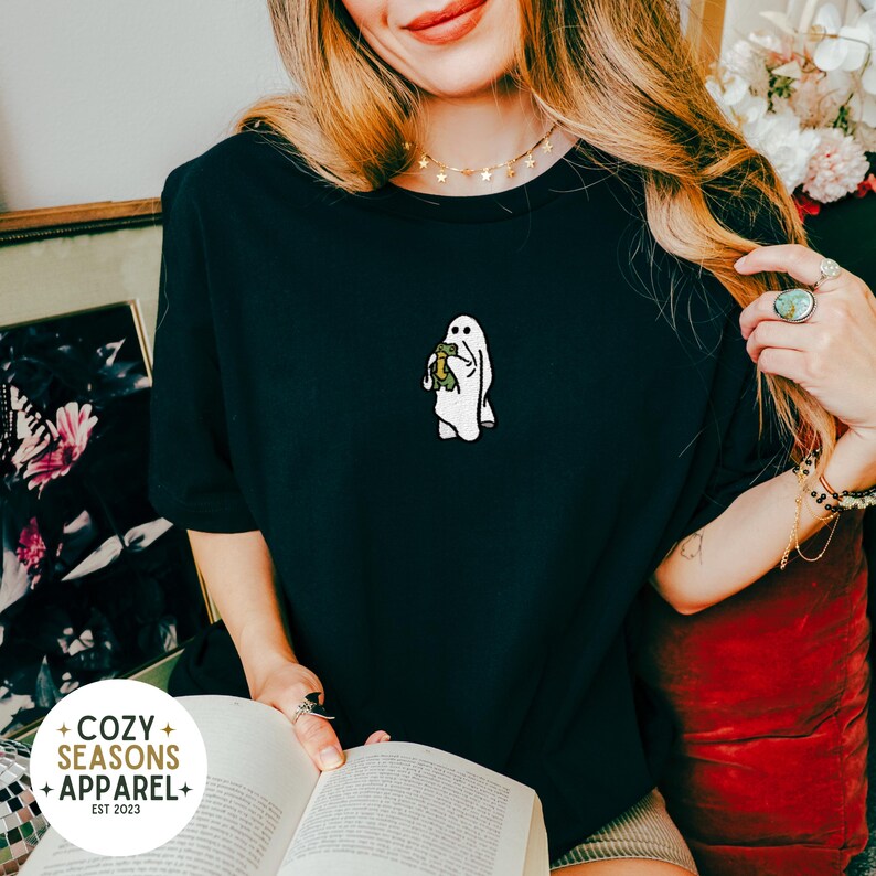 May include: A black t-shirt with a white ghost holding a green turtle embroidered on the front. The t-shirt is worn by a person with long blonde hair.