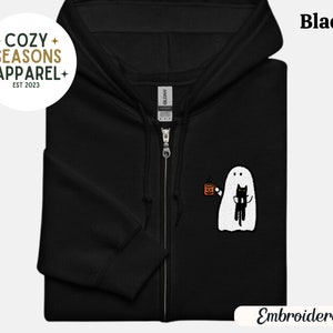 Peut inclure: Sweat-shirt à capuche noir zippé brodé avec un fantôme tenant un chat et une tasse de café. Le sweat-shirt porte le texte "Cozy Seasons Apparel Est 2023" sur le côté gauche et "Embroidered" en bas à droite.