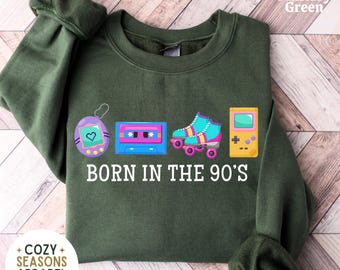 May include: Military green sweatshirt with a retro design featuring a Tamagotchi, cassette tape, roller skates, and a handheld game console. The text "BORN IN THE 90'S" is printed below the graphics. The sweatshirt has the "Cozy Seasons Apparel" logo.