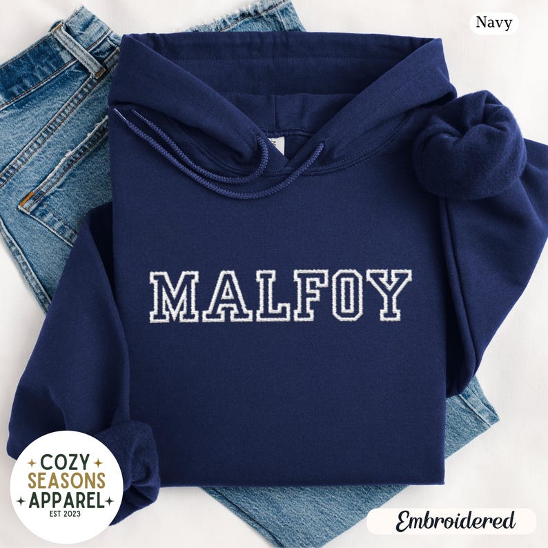 Peut inclure: Un sweat &agrave; capuche bleu marine avec le nom "MALFOY" brod&eacute; en blanc. Le sweat &agrave; capuche a une capuche &agrave; cordon et des manches longues. Les mots "Cozy Seasons Apparel" et "Embroidered" sont &eacute;galement visibles.