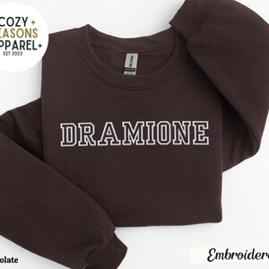 May include: A dark brown sweatshirt with the text "DRAMIONE" embroidered in white on the front. The sweatshirt is a crew neck style and has a relaxed fit.