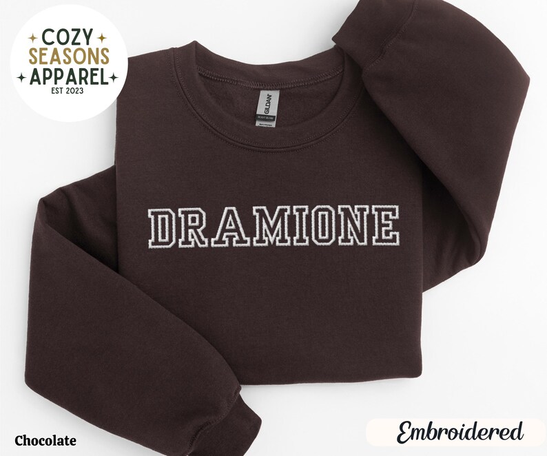 May include: A dark brown sweatshirt with the text "DRAMIONE" embroidered in white on the front. The sweatshirt is a crew neck style and has a relaxed fit.