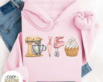 May include: Pink hoodie with a graphic of a mixer, piping bag, hand mixer, and cupcake. The text "Cozy Seasons Apparel" is on the bottom left corner of the hoodie.