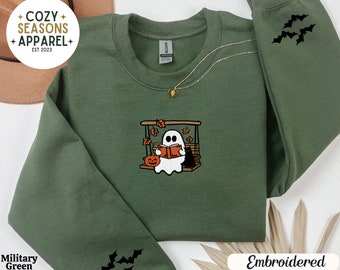 Può includere: Una felpa verde con un ricamo di un fantasma che legge un libro su un'altalena. Il fantasma è bianco e l'altalena è marrone. La felpa ha il testo "Cozy Seasons Apparel" e "Est 2023" sul davanti. Le maniche hanno pipistrelli ricamati.