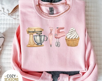 May include: Pink sweatshirt with a graphic design of a stand mixer, piping bag, whisk, and cupcake. The text "Cozy Seasons Apparel Est 2023" is on the bottom left corner of the image.