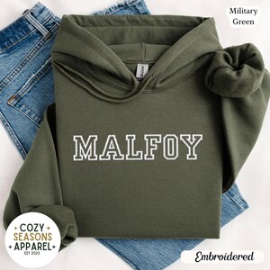 Peut inclure: Un sweat &agrave; capuche vert militaire avec le nom "MALFOY" brod&eacute; en blanc. Le sweat &agrave; capuche a une capuche &agrave; cordon et une poche avant. Les mots "Cozy Seasons Apparel" sont imprim&eacute;s sur un cercle blanc. Le sweat est pli&eacute; sur un jean bleu.