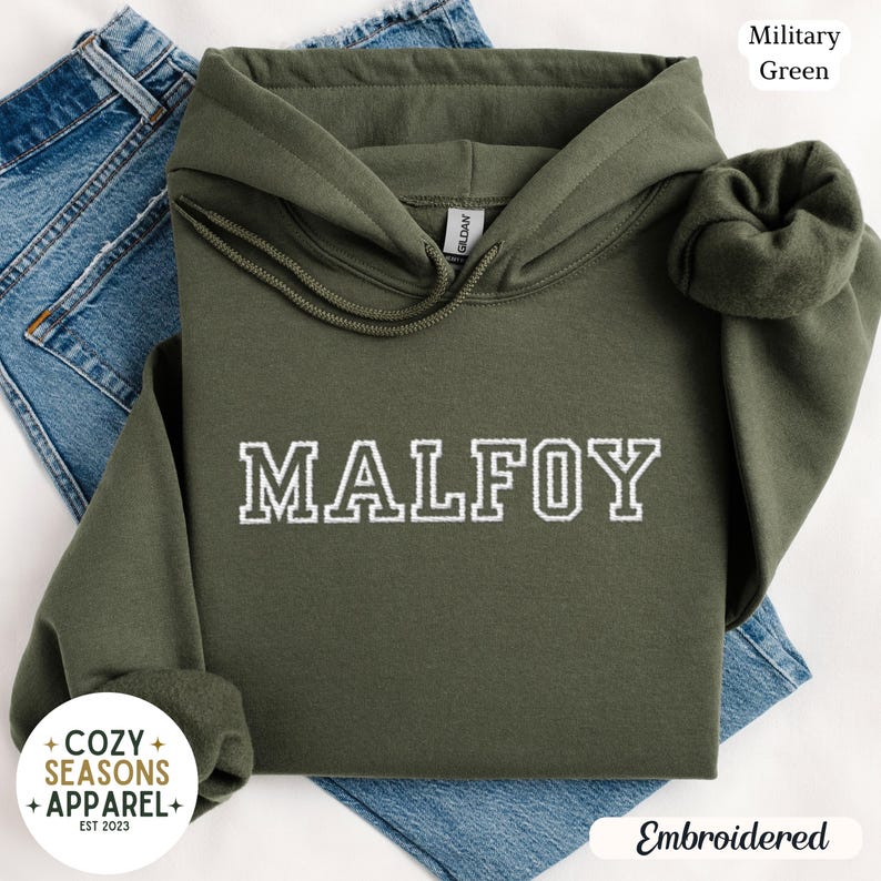 Peut inclure: Un sweat &agrave; capuche vert militaire avec le nom "MALFOY" brod&eacute; en blanc. Le sweat &agrave; capuche a une capuche &agrave; cordon et une poche avant. Les mots "Cozy Seasons Apparel" sont imprim&eacute;s sur un cercle blanc. Le sweat est pli&eacute; sur un jean bleu.