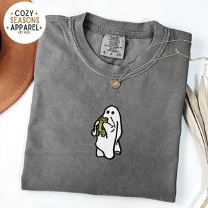 May include: A gray t-shirt with a white ghost holding a green frog embroidered on the front. The shirt has a tag that says "Comfort Colors" and "S - P - CH".