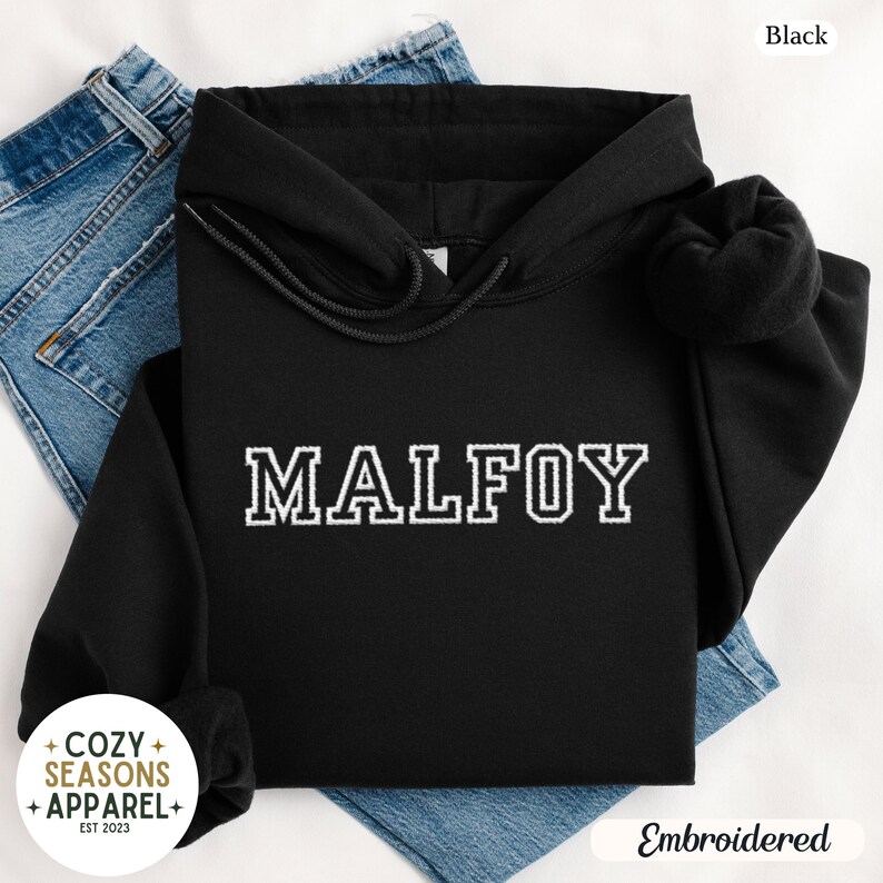 Peut inclure: Sweat &agrave; capuche noir avec le nom "MALFOY" brod&eacute; en lettres blanches. Le sweat-shirt a une capuche &agrave; cordon et des manches longues. Les mots "Cozy Seasons Apparel" sont imprim&eacute;s en bas &agrave; gauche.