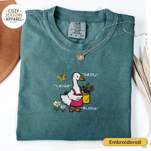 May include: Teal Comfort Colors t-shirt with an embroidered design featuring a goose wearing an apron and holding a yellow flower pot. The shirt has the words "LAUGH", "GROW", and "BLOOM" embroidered near the goose. A gold necklace is draped across the shirt.