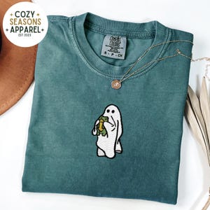 May include: A teal blue t-shirt with a white ghost holding a green frog embroidered on the front. The shirt has a tag that says "Comfort Colors" and "S - P - CH".