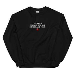 Op de afbeelding: Zwart sweatshirt met witte tekst die "Ik werd leraar voor het geld en de roem" luidt met een rode appelafbeelding.