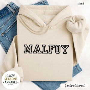 Peut inclure: Un sweat &agrave; capuche couleur sable avec le mot "MALFOY" brod&eacute; en lettres noires. Le sweat &agrave; capuche a une capuche &agrave; cordon et des manches longues. Le logo Cozy Seasons Apparel est visible en bas &agrave; gauche.
