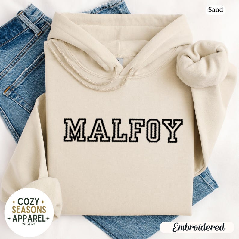 Peut inclure: Un sweat &agrave; capuche couleur sable avec le mot "MALFOY" brod&eacute; en lettres noires. Le sweat &agrave; capuche a une capuche &agrave; cordon et des manches longues. Le logo Cozy Seasons Apparel est visible en bas &agrave; gauche.