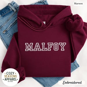 Peut inclure: Un sweat &agrave; capuche bordeaux avec le nom "MALFOY" brod&eacute; en blanc. Le sweat &agrave; capuche a une capuche &agrave; cordon et des manches longues. Les mots "Cozy Seasons Apparel" sont dans un logo circulaire en bas &agrave; gauche. Le mot "Embroidered" est en bas &agrave; droite.