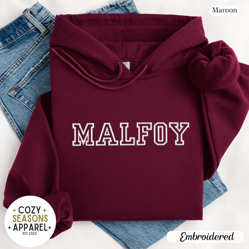 Peut inclure: Un sweat &agrave; capuche bordeaux avec le nom "MALFOY" brod&eacute; en blanc. Le sweat &agrave; capuche a une capuche &agrave; cordon et des manches longues. Les mots "Cozy Seasons Apparel" sont dans un logo circulaire en bas &agrave; gauche. Le mot "Embroidered" est en bas &agrave; droite.