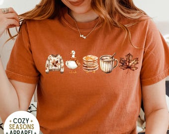 以下が含まれることがあります： 秋をテーマにしたイラストが並んだ、オレンジ色のTシャツ。秋の葉っぱのセーター、マフラーをしたアヒル、パンケーキ、ホットチョコレートのマグカップ、カエデの葉っぱなどが描かれています。Tシャツには「COZY SEASONS APPAREL + EST 2023」の文字が入っています。