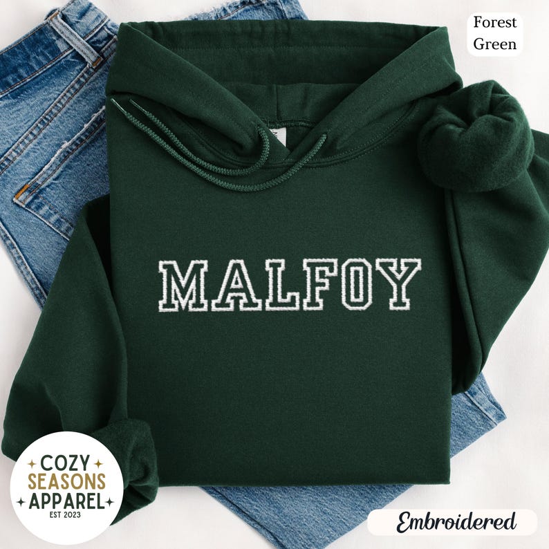 Peut inclure: Un sweat &agrave; capuche vert for&ecirc;t avec le nom "MALFOY" brod&eacute; en blanc. Le sweat &agrave; capuche a une capuche &agrave; cordon et des manches longues. Les mots "Cozy Seasons Apparel" sont imprim&eacute;s en bas &agrave; gauche.