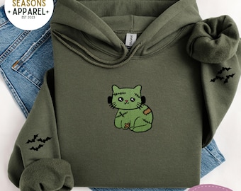 以下が含まれることがあります： フランケンシュタイン猫のデザインが刺繍されたオリーブグリーンのパーカー。猫は緑色で、黒いステッチとパッチが付いています。黒いコウモリが袖に刺繍されています。パーカーはブルージーンズの上に折りたたまれています。「Cozy Seasons Apparel」の文字が左上にあります。