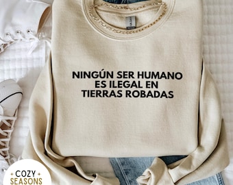 Può includere: Una felpa beige con la scritta "NINGÚN SER HUMANO ES ILEGAL EN TIERRAS ROBADAS" stampata in nero.