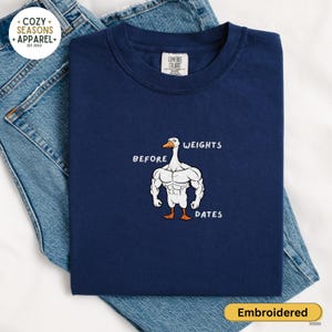May include: Navy blue sweatshirt with an embroidered design of a muscular goose. The text above the goose reads "WEIGHTS", and below it reads "DATES". The sweatshirt is displayed on a pair of blue jeans.