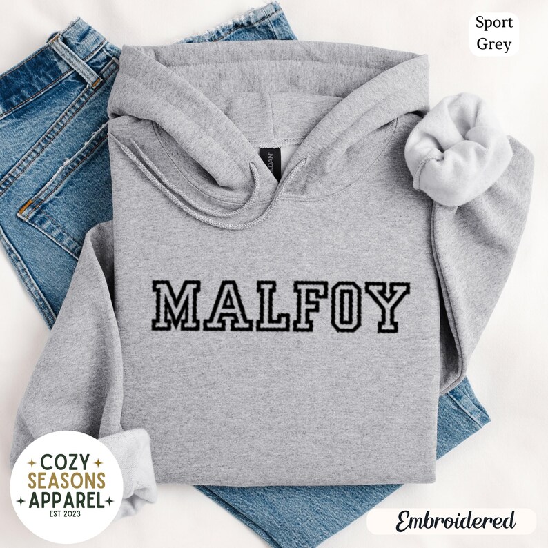 Peut inclure: Un sweat &agrave; capuche gris clair avec le nom "MALFOY" brod&eacute; en lettres noires. Le sweat &agrave; capuche a une capuche &agrave; cordon et un chouchou blanc. Les mots "Sport Grey" et "Embroidered" sont &eacute;galement visibles.