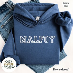 Peut inclure: Un sweat &agrave; capuche bleu indigo avec le mot "MALFOY" brod&eacute; en blanc. Le sweat &agrave; capuche a une capuche &agrave; cordon et est fait d'une mati&egrave;re douce. L'image montre &eacute;galement un jean bleu.