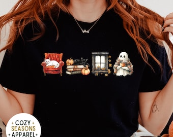 以下が含まれることがあります： 黒いTシャツ。秋のデザインで、赤い椅子に座る白い猫、カボチャと本の上にいる黒い猫、ろうそくのある窓、マグカップを持ったお化けが描かれています。「COZY SEASONS APPAREL EST 2023」の文字も表示されています。