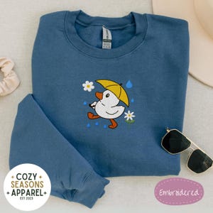 May include: A blue sweatshirt with a cute embroidered design of a duck wearing a yellow umbrella and walking in the rain. The design includes small white flowers and raindrops. The sweatshirt has the text "Cozy Seasons Apparel Est 2023" on the bottom left corner and "Embroidered" on the bottom right corner.