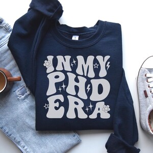 May include: Navy blue sweatshirt with the text "IN MY PHD ERA" in white, retro-style lettering. The design includes flower and star accents. The sweatshirt is paired with light-wash jeans, a coffee mug, and gray sneakers.