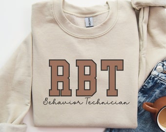 May include: A beige sweatshirt with the text "RBT Behaviour Technician" in brown letters with a black outline.