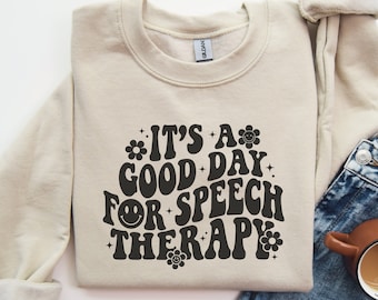 Puede incluir: Sudadera beige con la frase "IT'S A GOOD DAY FOR SPEECH THERAPY" en negro, con letras de estilo retro. El diseño incluye flores y una carita sonriente. La sudadera está doblada, con una parte de unos vaqueros a la vista.