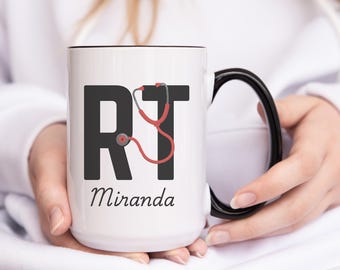 Puede incluir: Taza de cerámica blanca con asa y borde negros. La taza tiene un gráfico negro y rojo de las letras "RT" con un estetoscopio envuelto alrededor de las letras. El texto "Miranda" está impreso debajo del gráfico.