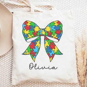 May include: A white tote bag with a colorful puzzle piece bow and the name "Olivia" printed on the front.
