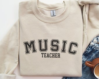May include: A beige sweatshirt with the text "MUSIC TEACHER" in a black varsity-style font.